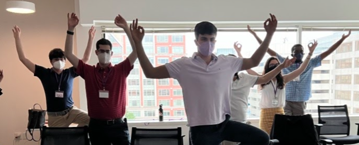 This is a photograph of the Summer 2022 interns practicing yoga. They are all standing on one leg with their hands above their heads.
